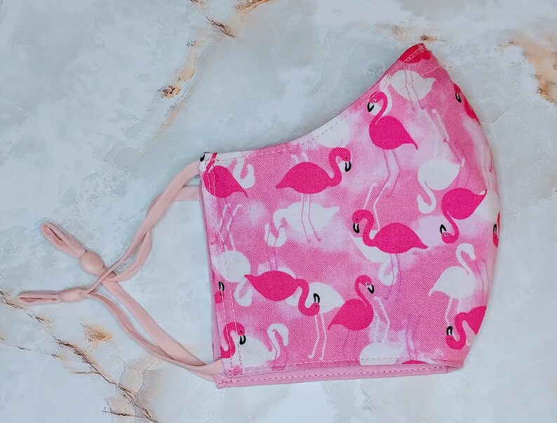 May include: Pink face mask with a flamingo print. The mask has adjustable ear loops and a pink and white pattern.