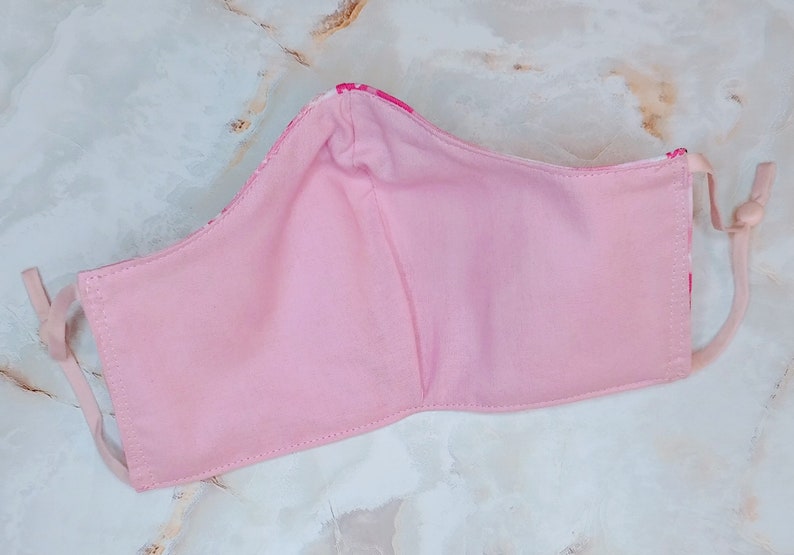 May include: Pink fabric face mask with white floral print. The mask has two ties for the ears.