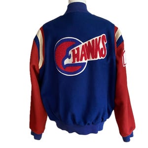 May include: A blue varsity jacket with red sleeves and cream-colored shoulder accents. The back features a large circular patch with a stylized eagle head profile and the text HAWKS in red letters. The jacket has a ribbed collar and cuffs.