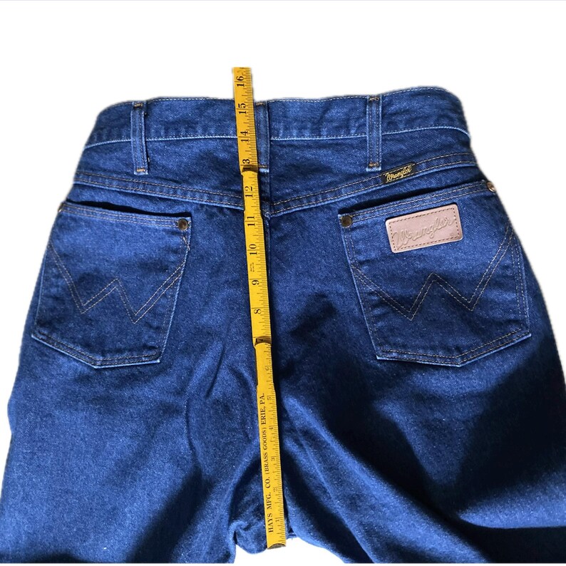 May include: Dark blue Wrangler jeans with a yellow measuring tape showing a 13-inch inseam. The jeans have classic back pockets with the Wrangler logo and a leather patch. The jeans are made of denim.