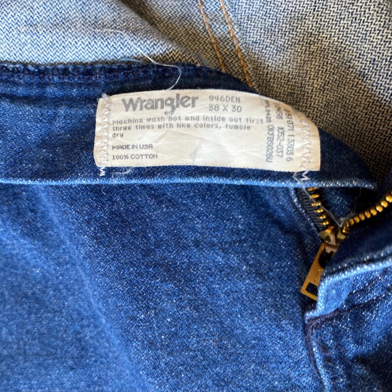 May include: Close-up of a pair of blue denim Wrangler jeans. The tag reads "Wrangler 946DEN 38 x 30" and "100% Cotton". The jeans have a zipper and a button closure.