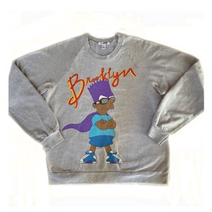 May include: A gray sweatshirt featuring a cartoon character with purple hair, sunglasses, and a cape. The word "Brooklyn" is written in red and yellow script above the character. The text "WE GO HARD" is also visible.