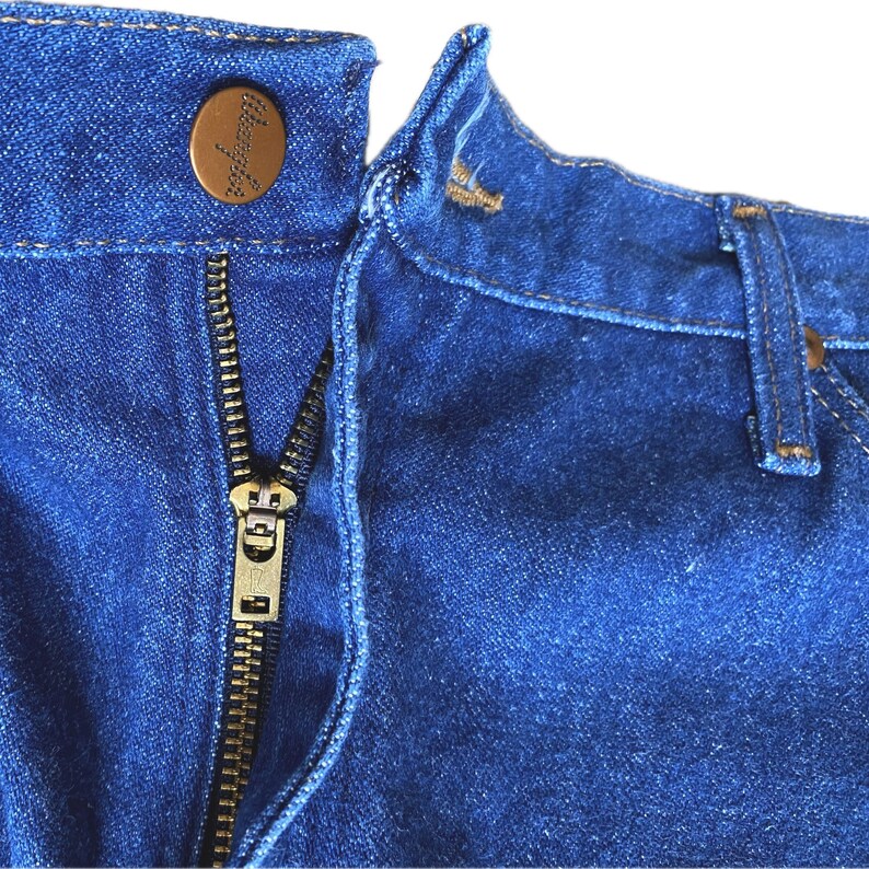 May include: Close-up of blue denim jeans with a brass zipper and a button closure. The button has the Wrangler logo. The jeans are a classic style, with a focus on the details of the zipper and button.