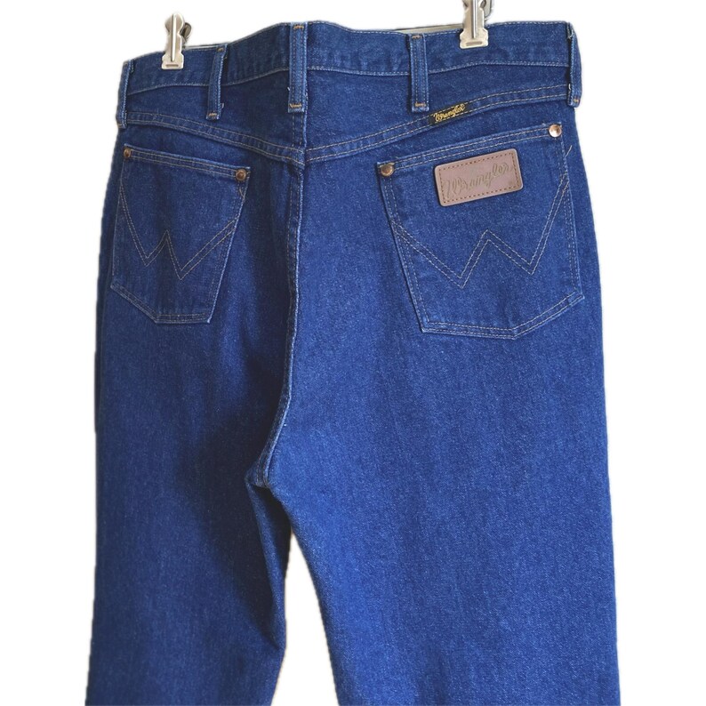 May include: Back view of blue denim jeans with the Wrangler logo on the back pocket. The jeans have a classic five-pocket design, with the signature Wrangler stitching on the back pockets. The jeans are hanging on a hanger.
