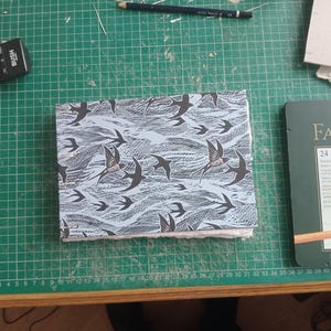 May include: A handmade journal with a swallows pattern on the cover, set against a light blue and white backdrop. The journal is on a green cutting mat, with art supplies nearby. The cover design features swallows.