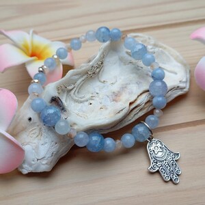 Può includere: Un braccialetto di pietre di acquamarina blu con un ciondolo a mano di Hamsa argentato. Il braccialetto è su una conchiglia bianca con fiori rosa.