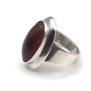 May include: A sterling silver ring featuring a smooth, oval-shaped dark red stone set in a polished metal bezel. The interior of the band is engraved with the text STERLING, DENMARK, and FROM. The ring has a wide, solid band design.