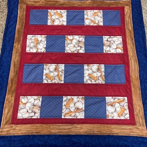 May include: A blue and red baseball-themed quilt with a wooden frame. The quilt features a pattern of baseballs and bats on a blue background.