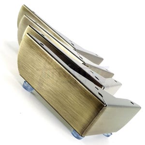 May include: A set of four metal furniture legs in varying shades of gold and silver. Each leg has a rectangular body with a curved front and is mounted on a clear, rounded foot. The legs are angled and appear to be new.