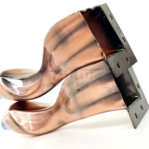 Puede incluir: Dos patas de muebles de metal de color cobre con un diseño curvo. Las patas tienen un soporte de metal negro con agujeros para montaje.