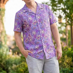 May include: A purple button-down shirt with a floral pattern, paired with gray shorts. The shirt features a vibrant design with shades of purple, pink, and yellow. The image is set outdoors with a castle in the background.