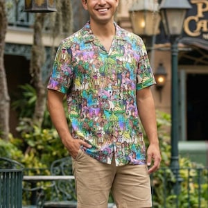 May include: A colorful short-sleeve button-down shirt with a tropical print, paired with khaki shorts and white sneakers. The shirt features a vibrant mix of green, blue, purple, and yellow. The background includes a sign that says "Pirates of the Caribbean".