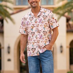 May include: A short-sleeved button-down shirt in a light pink hue featuring a repeating pattern of cartoon characters wearing sombreros and playing guitars, paired with blue denim shorts and white sneakers.