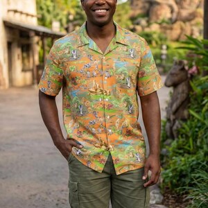 May include: Orange button-down shirt with a colorful jungle-themed pattern, paired with olive green cargo shorts. The shirt features short sleeves and a classic collar. The shorts have multiple pockets. The person is wearing white sneakers.