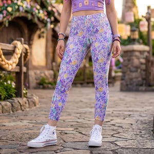 Peut inclure: Une femme porte un haut court lavande et des leggings à motifs floraux et de châteaux. Elle porte des baskets montantes blanches. En arrière-plan, un bâtiment de conte de fées et une enseigne "Tangled".