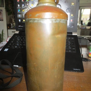 May include: A vintage copper water bottle with a brass lid and handle. The bottle has a worn, aged patina and is slightly dented.
