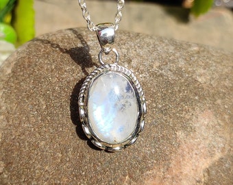 May include: An oval moonstone pendant set in a polished silver frame with a twisted rope border detail. The gemstone displays a translucent white appearance with subtle blue iridescence. The pendant hangs from a delicate silver chain necklace.