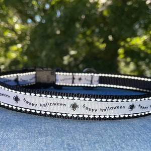 May include: A black and white dog collar with a reflective strip and the text "happy halloween" repeated along the collar. The collar has a black buckle and a white background with black spider silhouettes.