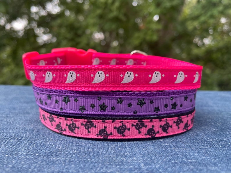 May include: A set of three dog collars with Halloween themes. The top collar is pink with white ghost silhouettes. The middle collar is purple with black glitter stars. The bottom collar is pink with black skull and crossbones.