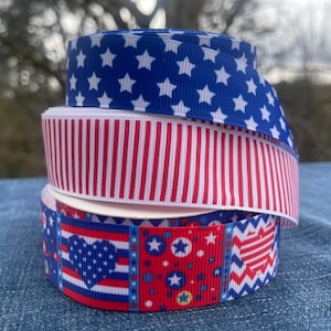 May include: Three spools of patriotic ribbon in red, white, and blue. The top spool features a blue background with white stars. The middle spool has red and white stripes. The bottom spool has a heart and star design.