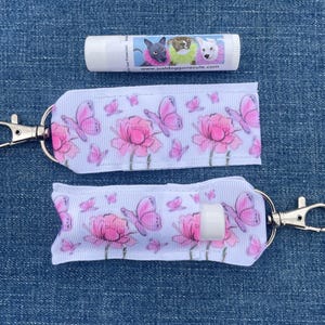 May include: Two white lip balm holders with a pink floral and butterfly pattern, each with a silver-colored clasp. A tube of lip balm with dog illustrations is above the holders. The website address "justgonecute.com" is visible on the lip balm.