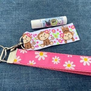 May include: Pink and white daisy patterned key chain with a silver key ring and a white lip balm with the text "Just Dog Smile Love". The lip balm has a picture of two dogs on it. The key chain also has a white ribbon with pink polka dots and three monkeys on it.