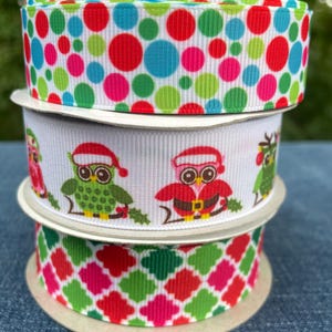 May include: Three spools of decorative ribbon. The top spool features a white background with large, colorful polka dots in red, green, blue, and pink. The middle spool has a white background with owl designs wearing Santa hats. The bottom spool has a green, pink, and red diamond pattern.