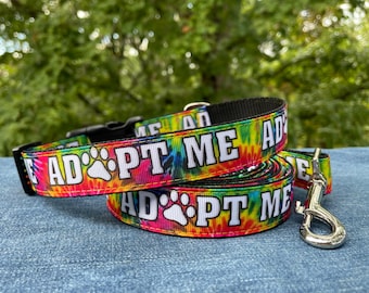 May include: A colorful tie-dye dog collar and leash set with the text "Adopt Me" in white lettering. The collar and leash are both decorated with a paw print.