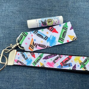 May include: A white key chain with a black border and a colorful crayon pattern. The keychain has a silver key ring and a silver clip. The keychain is paired with a white lip balm tube with a colorful dog pattern and the text "Just Did Gone Cute".