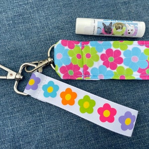 May include: A white keychain with a floral design and a silver clip. The keychain has a small pocket for a lip balm tube. The lip balm tube is white with a blue and white label that says "Just Dog Gone Cute".