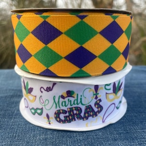 May include: Two spools of decorative ribbon. The top spool features a harlequin pattern in gold, green, and purple. The bottom spool is white with the words "Mardi Gras" and festive graphics in green, purple, and gold.