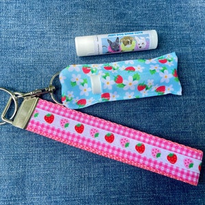 May include: A pink and white gingham ribbon keychain with a strawberry print. The keychain has a silver key ring and a blue and white lip balm tube with a strawberry print and the text "Just dog gone cute".