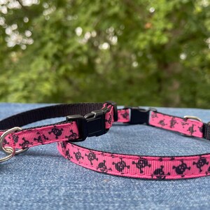 May include: Pink cat collar with a black skull pattern and a silver bell. The collar is adjustable and has a buckle closure.