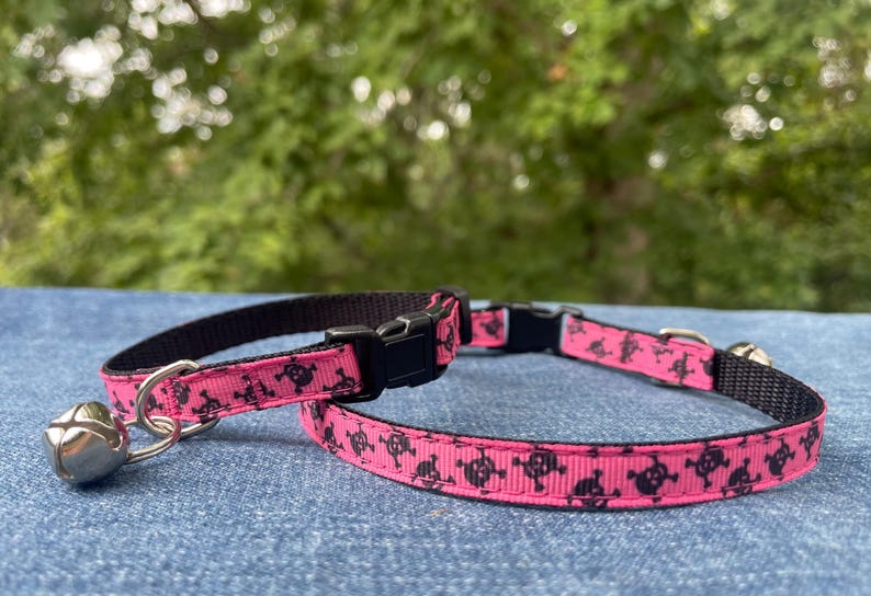 May include: Pink cat collar with a black skull pattern and a silver bell. The collar is adjustable and has a buckle closure.