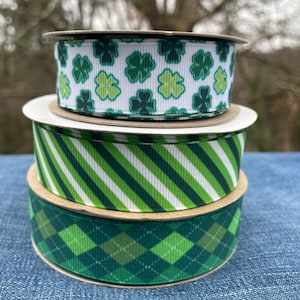 May include: Three spools of ribbon stacked on top of each other. The top spool has a white background with green shamrocks. The middle spool has green and white diagonal stripes. The bottom spool has a green plaid pattern.