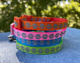 May include: Four cat collars with paw print designs. The collars are layered on top of each other, with the top collar being orange, followed by pink, blue, and green. The paw prints are silver.