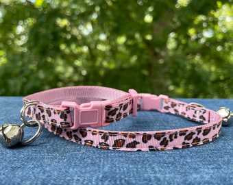 May include: Pink cat collar with a leopard print pattern and silver bells. The collar has an adjustable buckle.