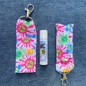 May include: Two white fabric keychains with a pink, green, and blue paint splatter design. Each keychain has a silver metal clasp and a silver metal ring. A white lip balm tube with a dog graphic and the text "Just Dog Gone Cute" is in the center of the image.