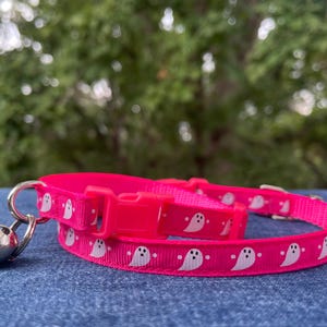 May include: A pink cat collar with a white ghost pattern and a silver bell. The collar is made of a soft, comfortable material and is adjustable to fit most cats.