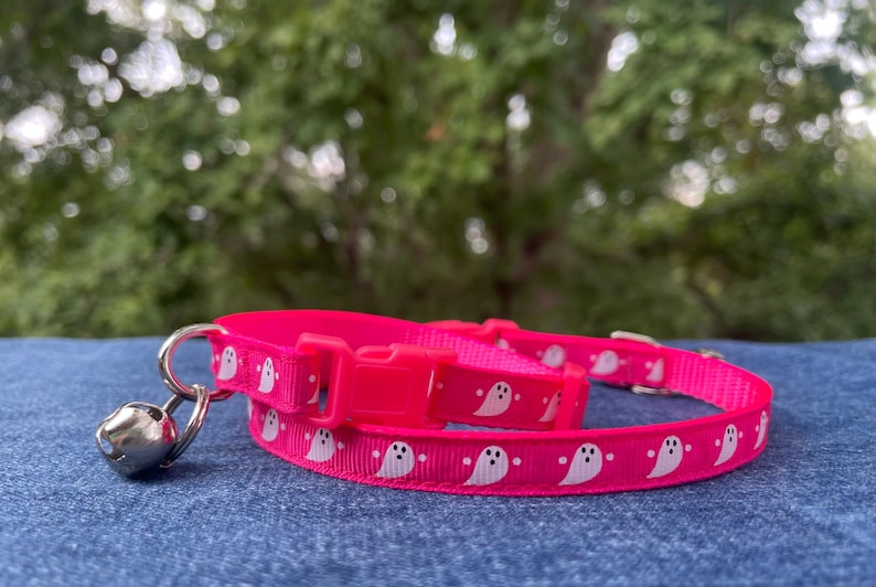 May include: A pink cat collar with a white ghost pattern and a silver bell. The collar is made of a soft, comfortable material and is adjustable to fit most cats.