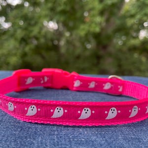 May include: A pink dog collar with a white ghost pattern. The collar is made of nylon and has a plastic buckle.