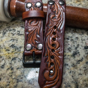 May include: Two brown leather watch straps with intricate floral designs and silver accents. The straps have a buckle fastening.