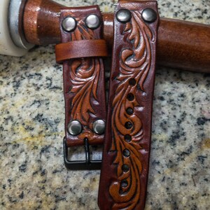 May include: Two brown leather watch straps with intricate floral designs and silver metal accents. The straps have a buckle fastening.