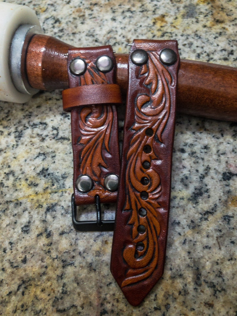 May include: Two brown leather watch straps with intricate floral designs and silver metal accents. The straps have a buckle fastening.