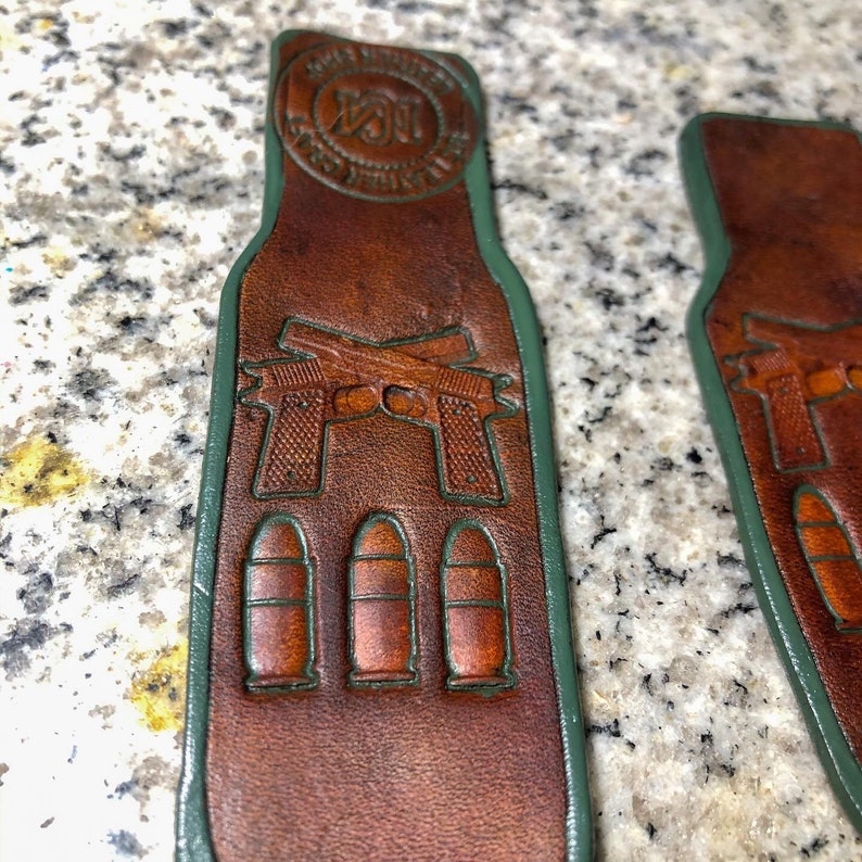 May include: Two brown leather gun belt loops with green stitching. The loops feature a design of two crossed pistols and three bullets. The loops have a circular logo with the text "Sons of Liberty" and a stylized "N" in the center.
