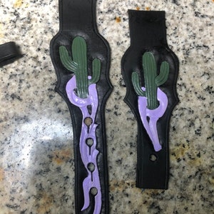 May include: Two black leather guitar straps with a purple and green cactus design. The straps have a decorative design with holes.