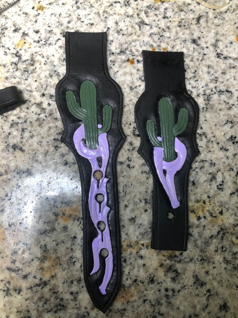May include: Two black leather guitar straps with a purple and green cactus design. The straps have a decorative design with holes.