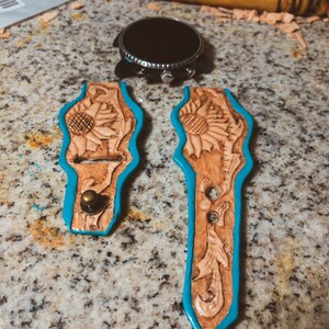 May include: Two unfinished leather watch straps with a sunflower design. The straps are trimmed in turquoise blue and have a brass button on one end.