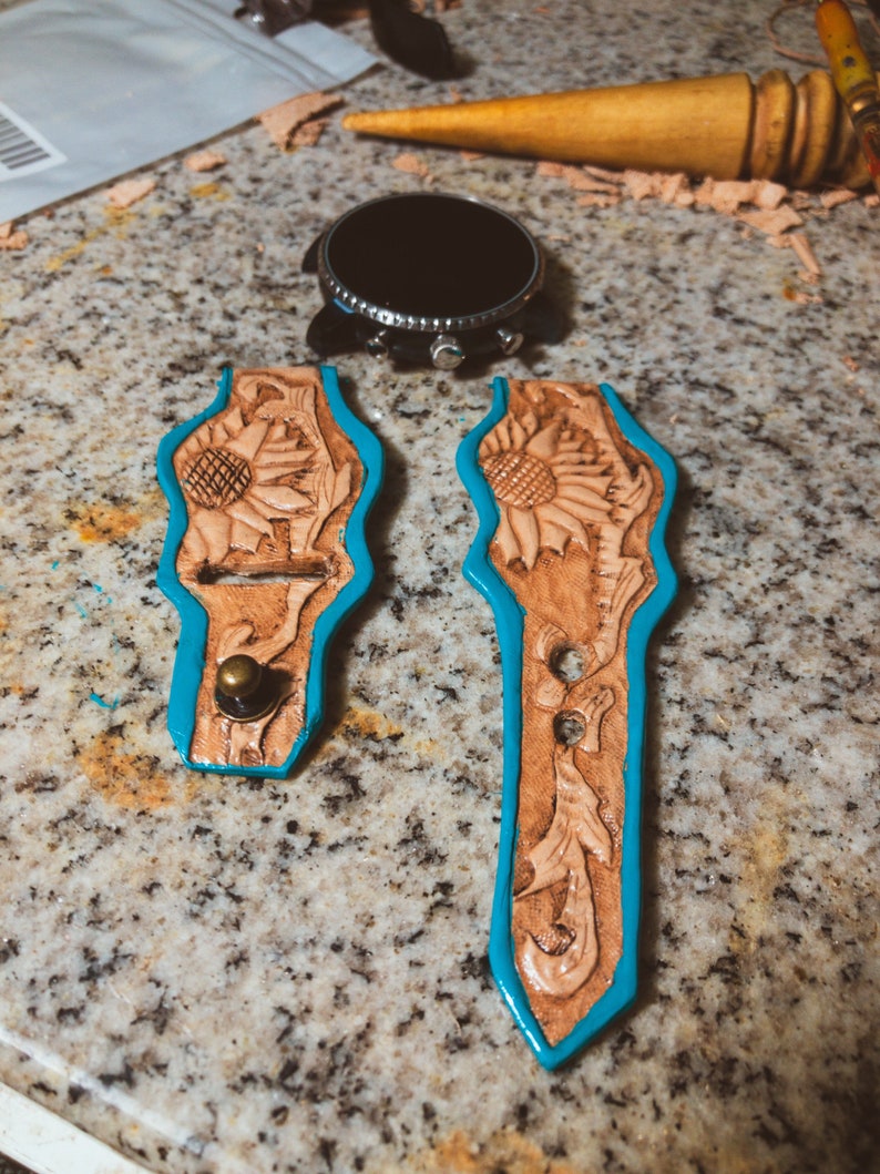 May include: Two unfinished leather watch straps with a sunflower design. The straps are trimmed in turquoise blue and have a brass button on one end.