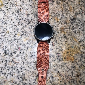 May include: A brown leather watch strap with a sunflower design. The strap is attached to a black smartwatch with a round face.